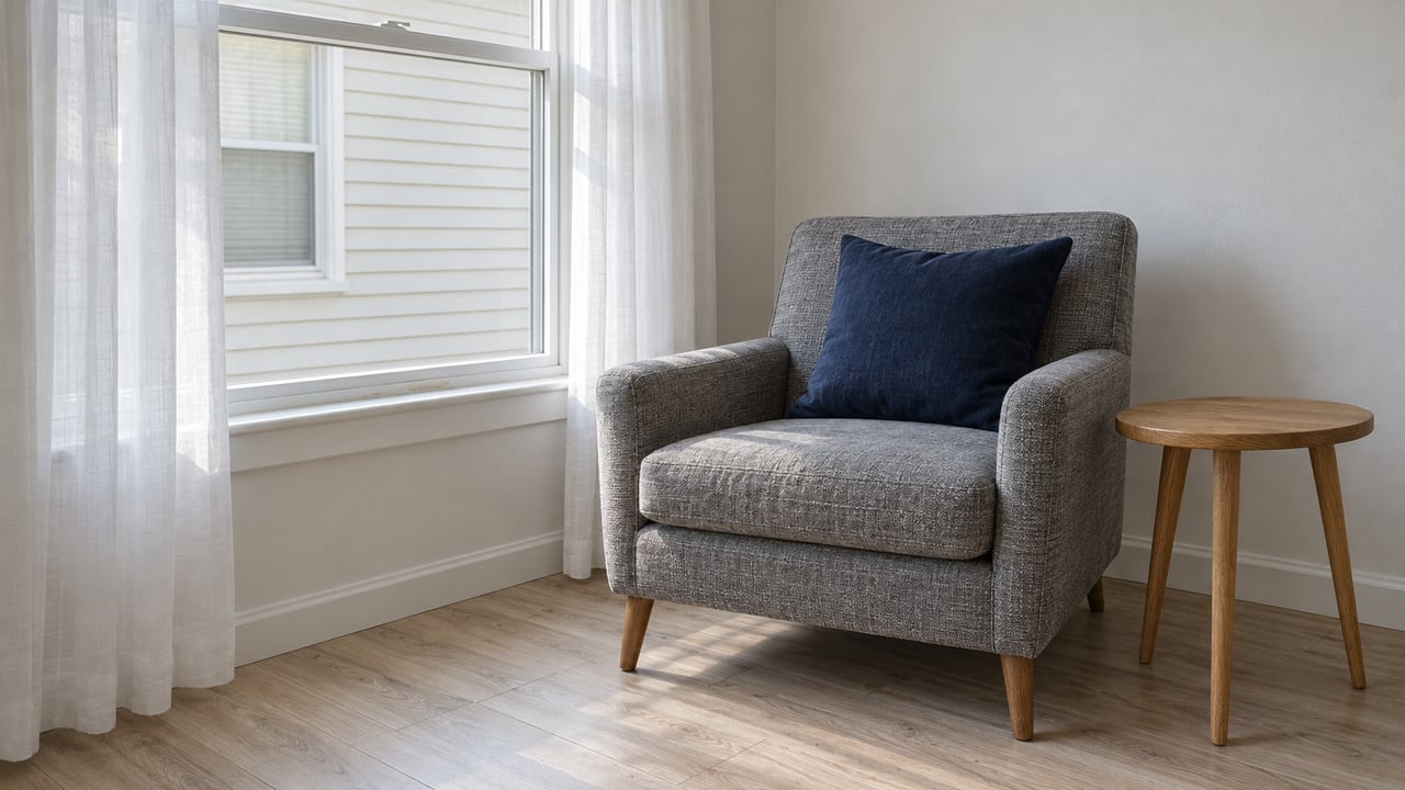 Soft daylight falls through sheer curtains onto a gray armchair and oak side table in a modest living room with pale flooring.