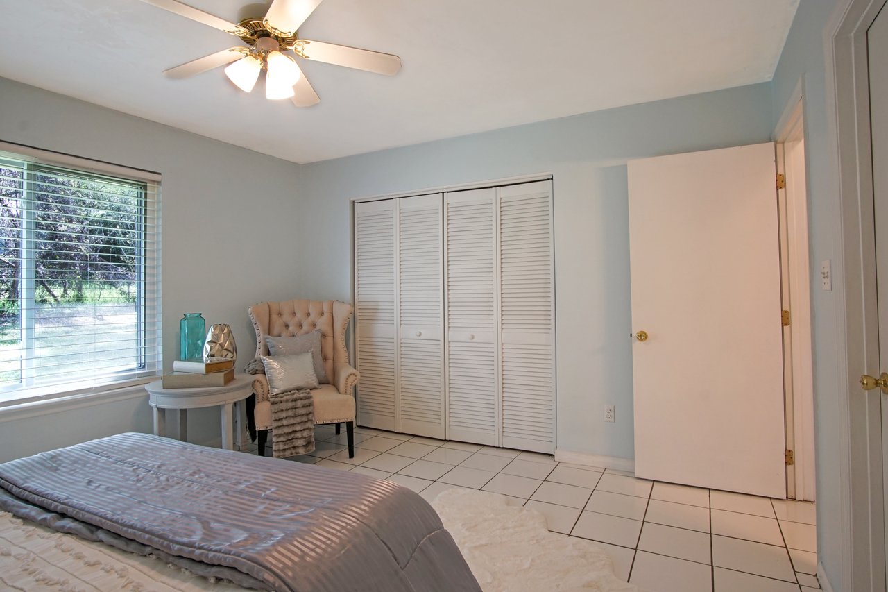 A serene bedroom with light blue walls, white tiles, and a bed with gray bedding. A cozy armchair, side table, and fan enhance the calm atmosphere.