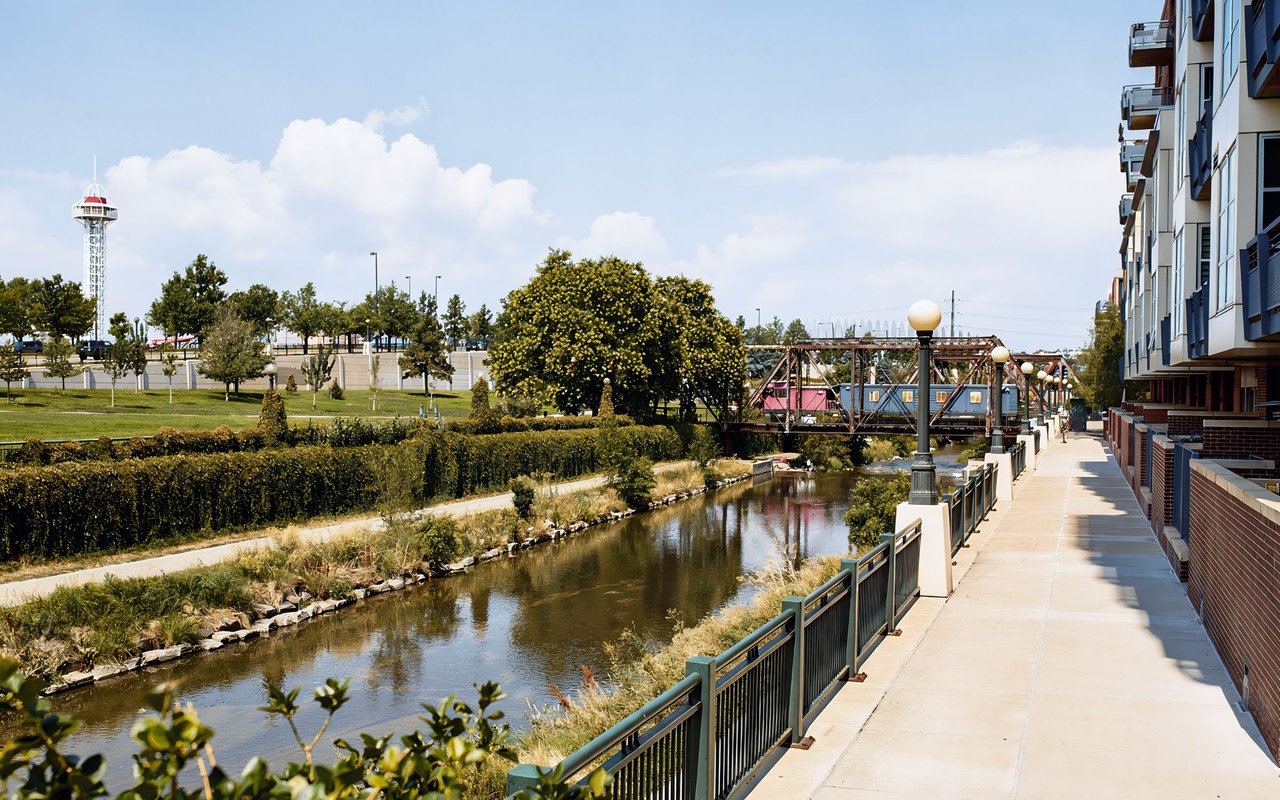 Urban creek beside a paved pedestrian path, with a metal bridge and modern apartment building.