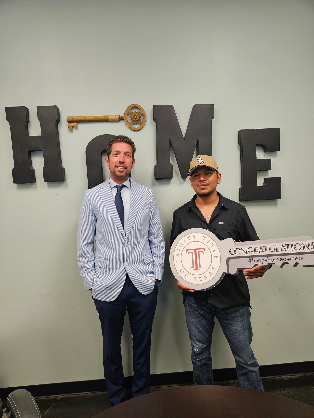 Two men pose with a large congratulatory key sign beneath the word HOME.