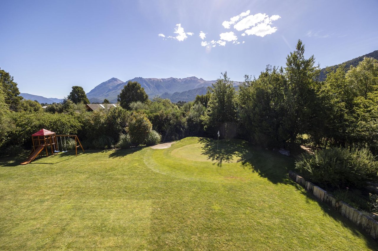 Landscaped backyard with a putting green and playset, backed by trees and mountain peaks.