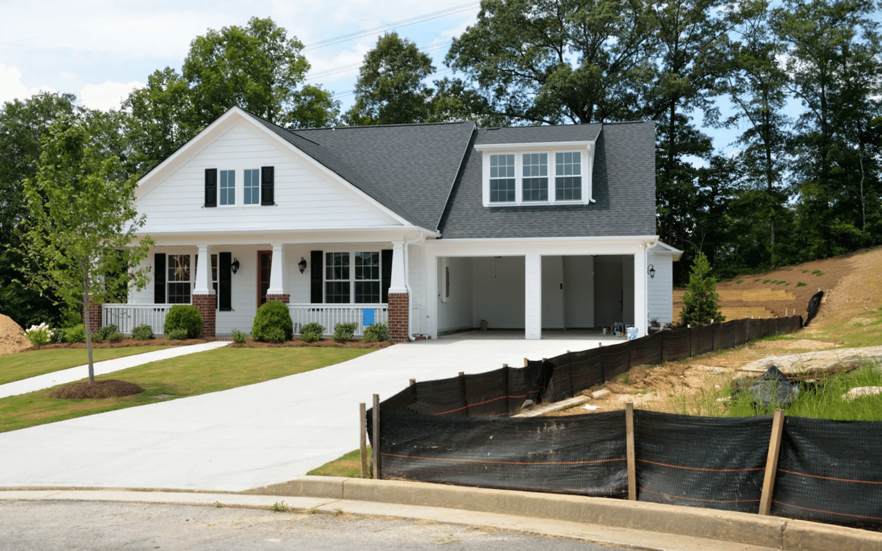 White two-story house with a covered porch, attached double garage, and construction fencing along the driveway.