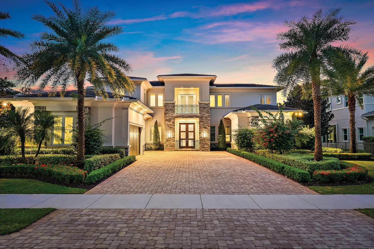 Two-story luxury home with stone accents, palm trees, and a wide paver driveway at dusk.