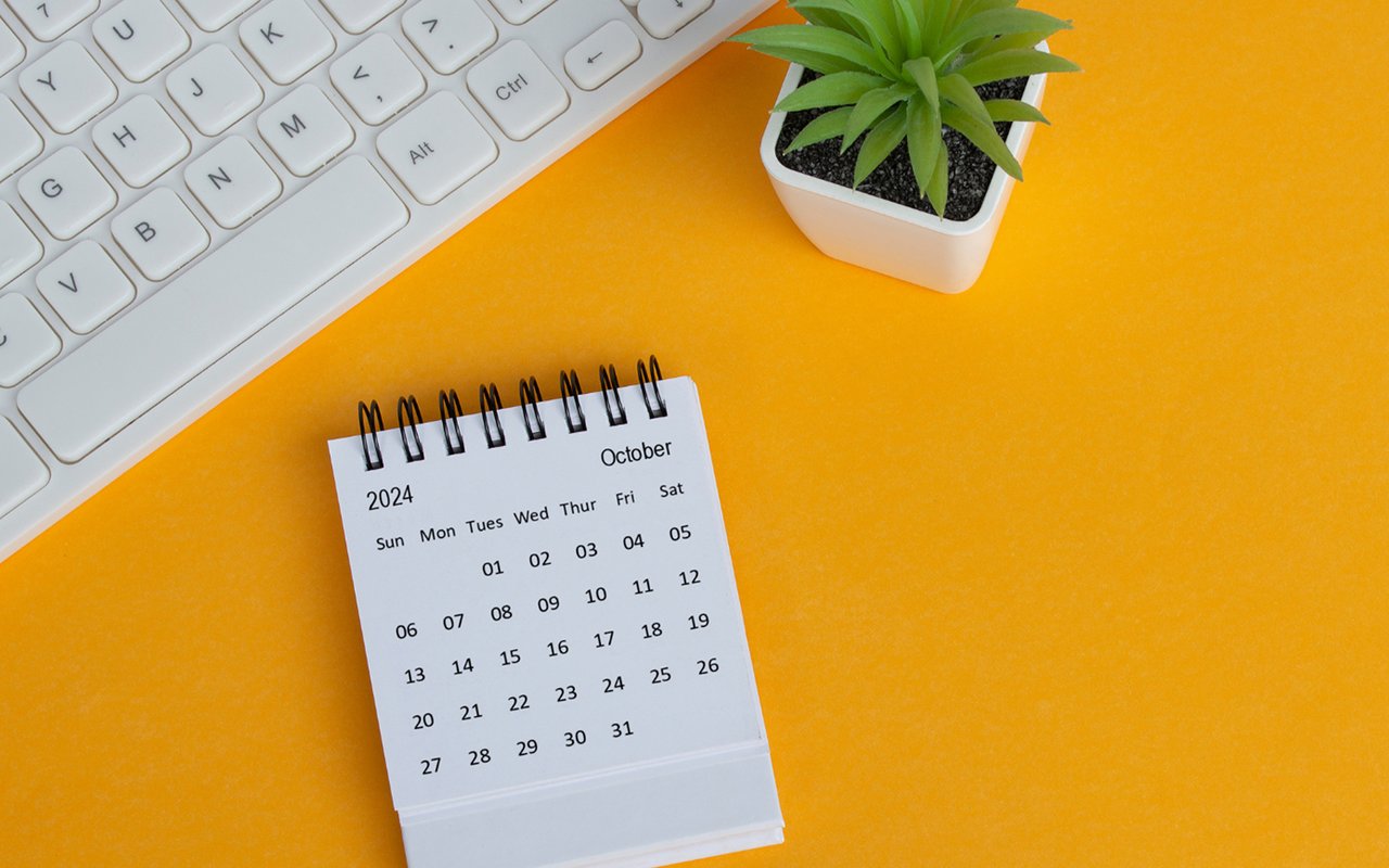 October 2024 desk calendar beside a white keyboard and small potted plant on an orange surface.