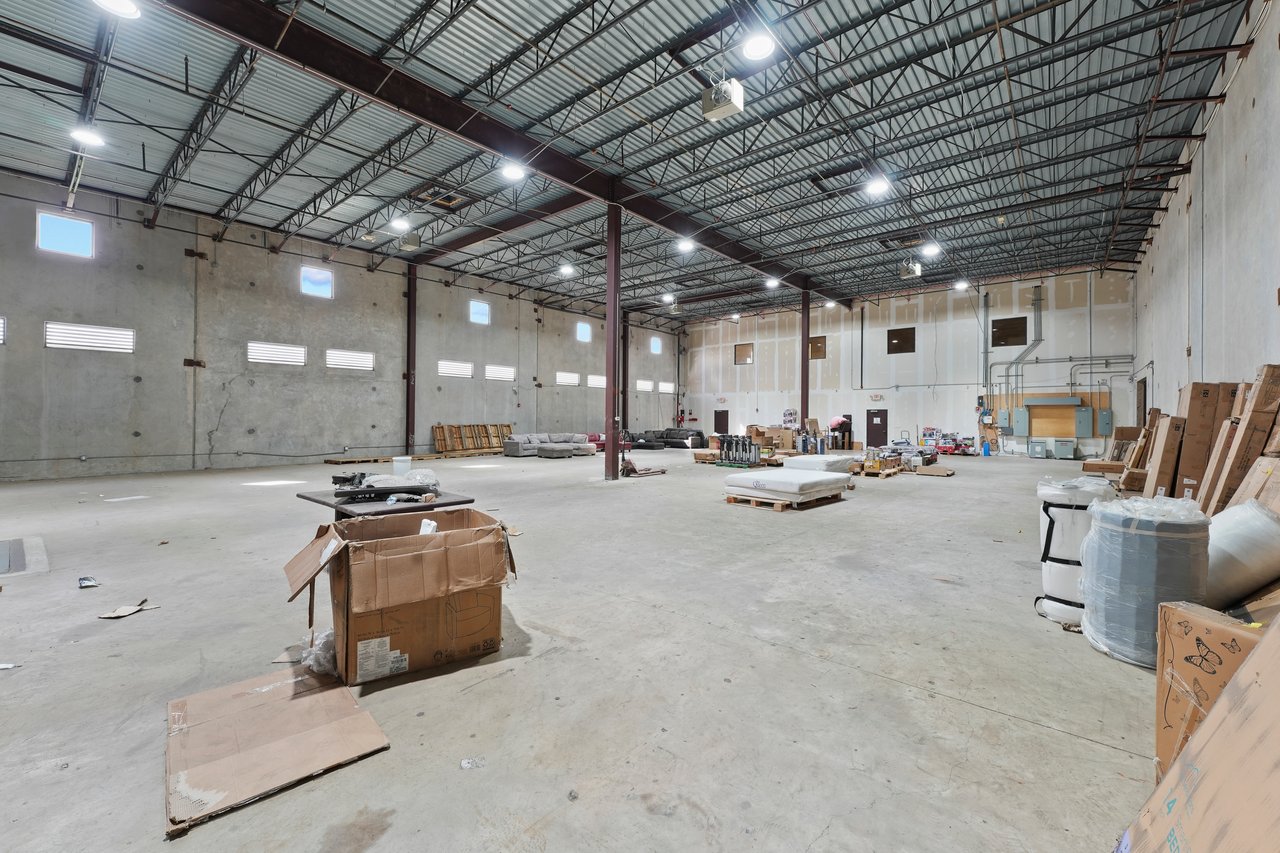Large warehouse interior with exposed steel roof trusses, concrete floor, and scattered boxes and equipment.