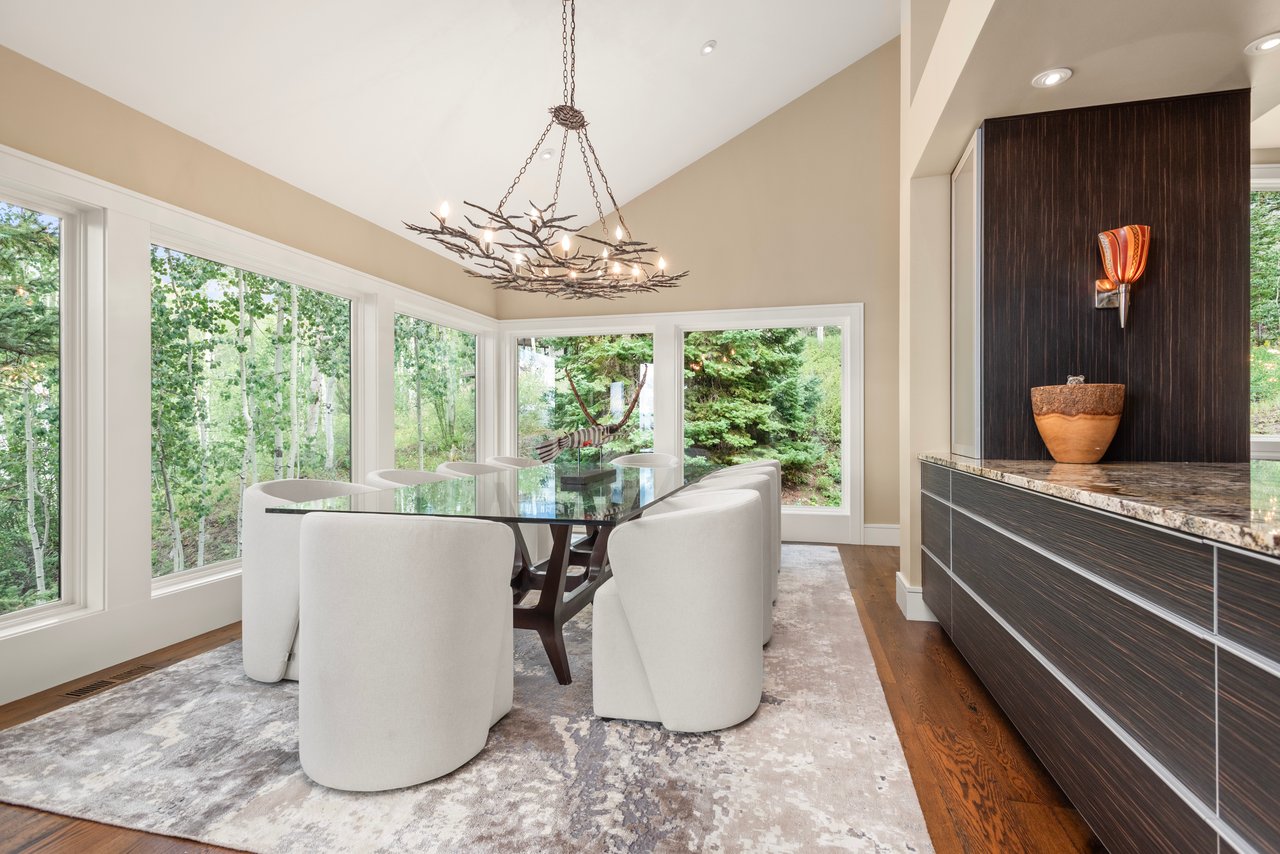 Dining room with a branch chandelier, glass table, white chairs, and large windows overlooking trees.