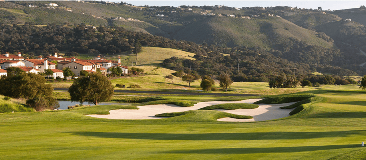 Golf course with a broad sand bunker, hillside homes, and wooded hills in warm light.