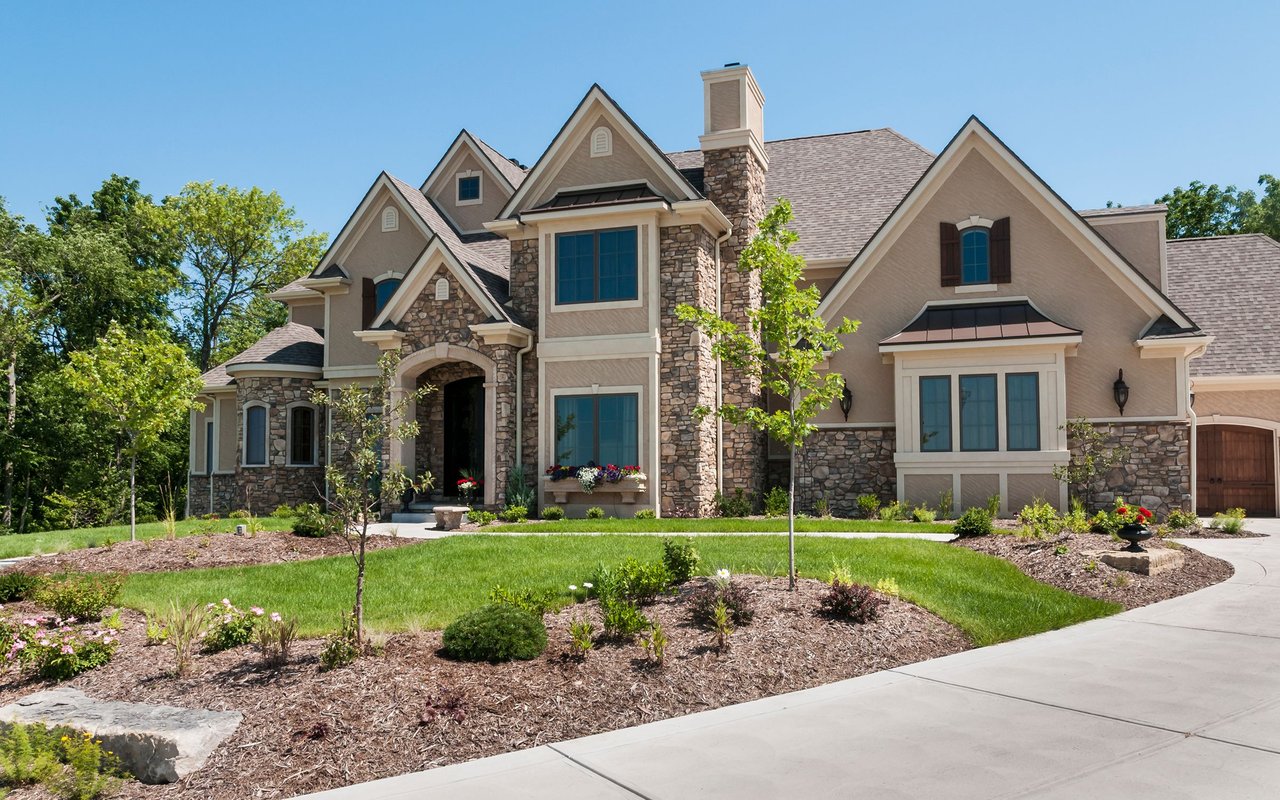 Large stone-and-tan home with multiple gables, landscaped gardens, and a curved driveway.