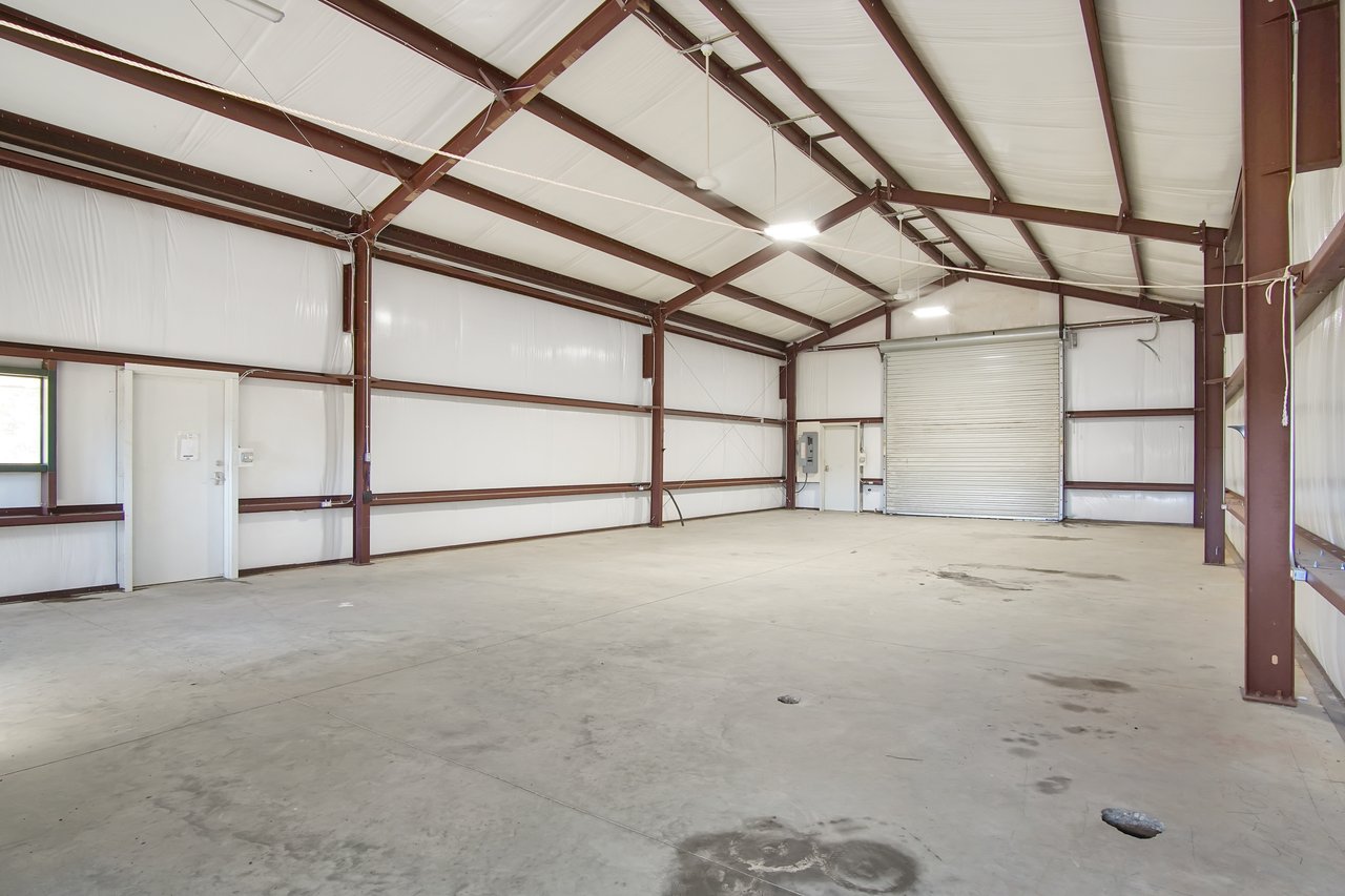 Empty industrial warehouse interior with high metal beams, white insulation, concrete floor, a small door on the left, and a large rolling door.