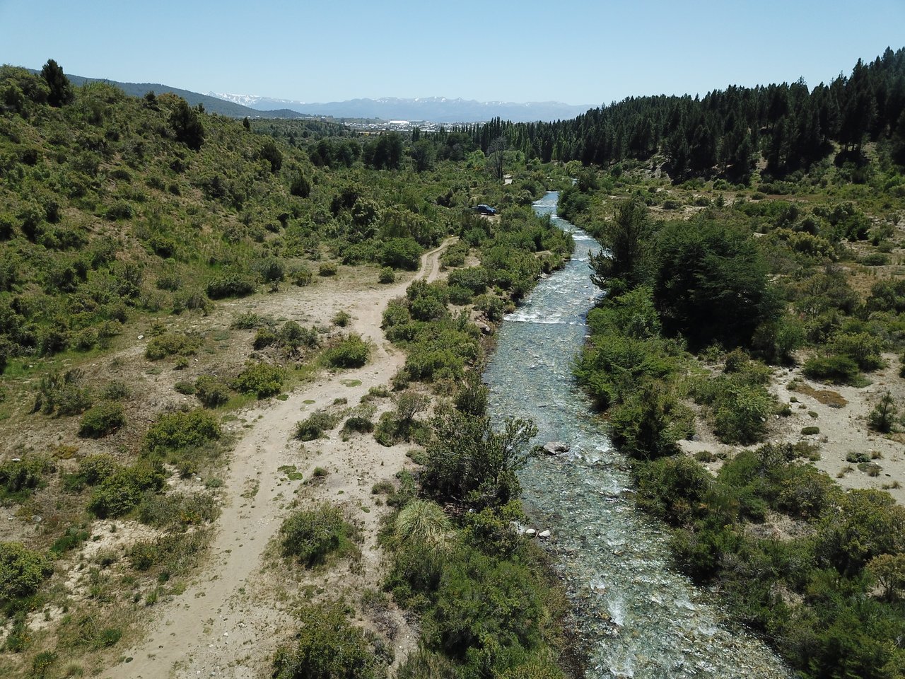 Vacant land beside a winding river, with forested hills and distant snow-capped mountains.
