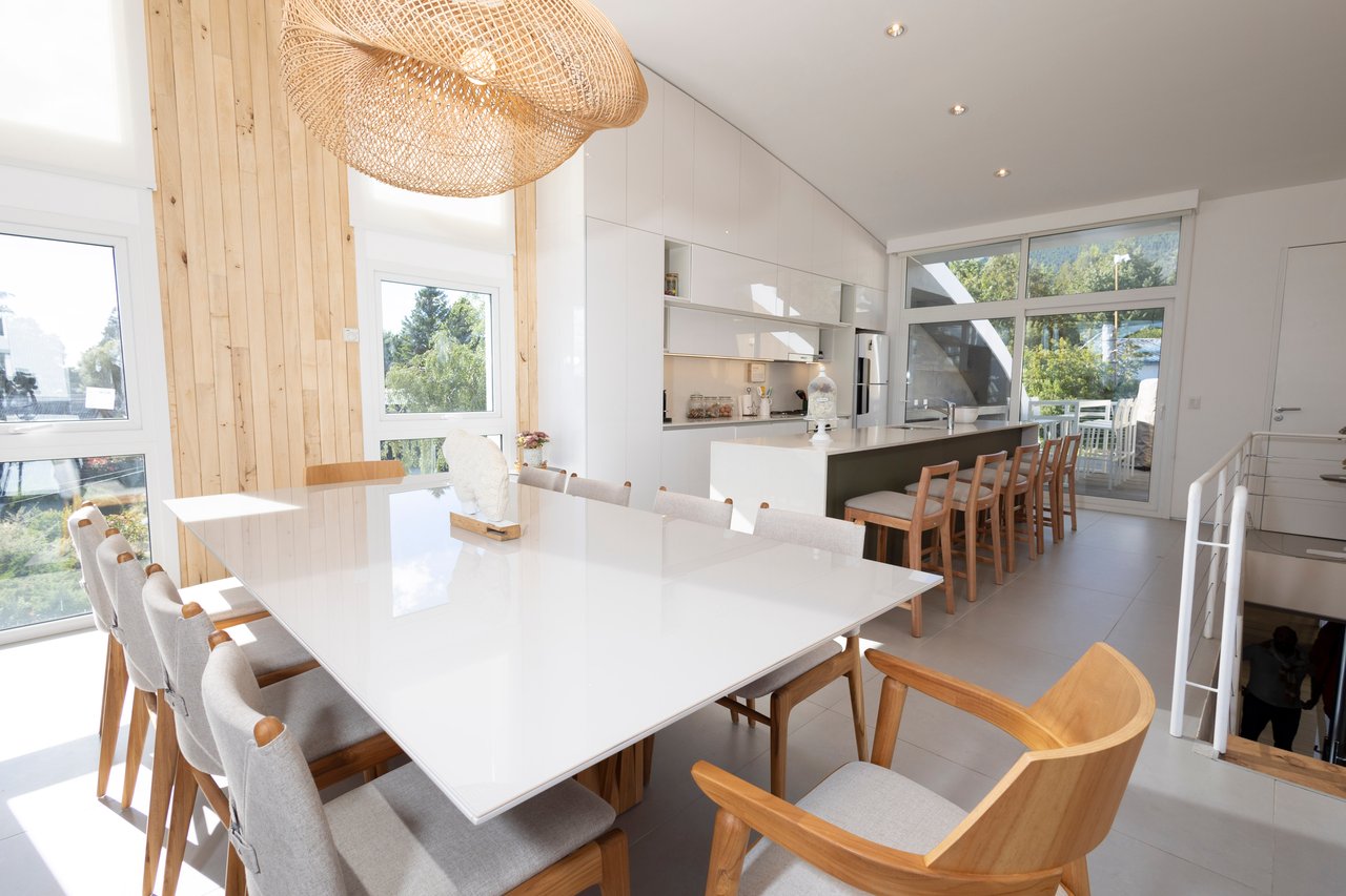 Open-plan dining room with a large white table, kitchen island, and tall windows overlooking trees.