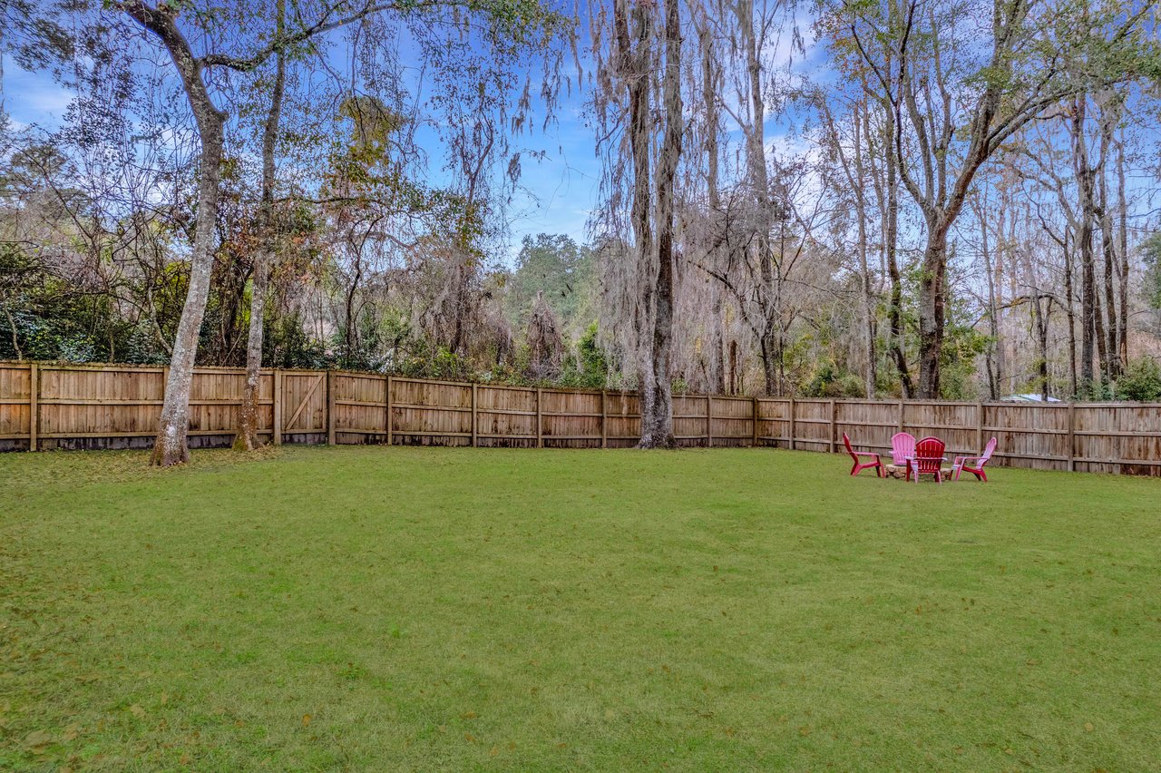 A grassy backyard enclosed by a wooden fence, featuring tall trees with hanging moss. Four red Adirondack chairs encircle a fire pit, under a clear sky.