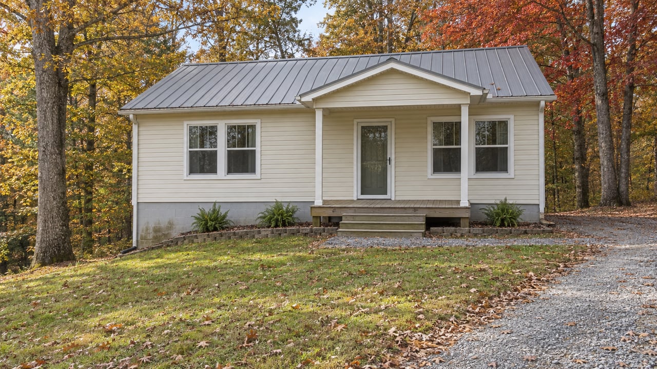 Cream-sided cottage with a gray metal roof, small wooden porch, sloping lawn and gravel drive beneath autumn oak and maple trees.