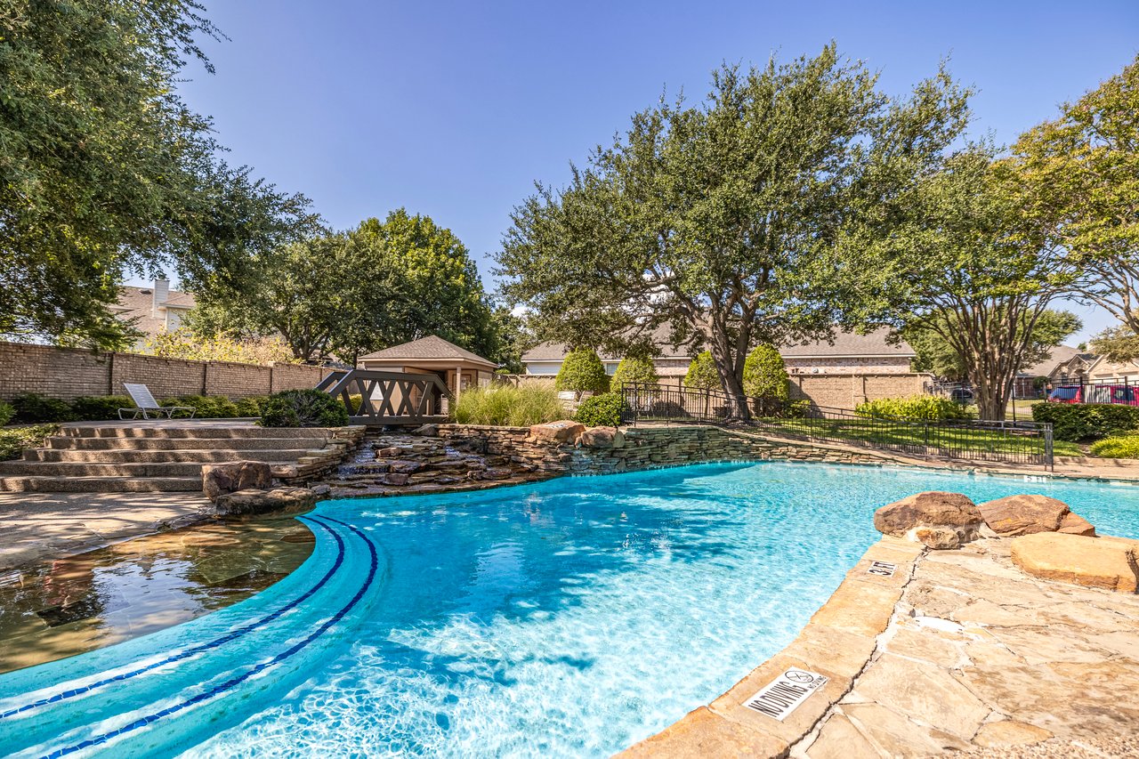 A sparkling blue pool with a wooden trestle bridge and waterfall. This is the smallest and most intimate of the Woobridge neighborhood pools.