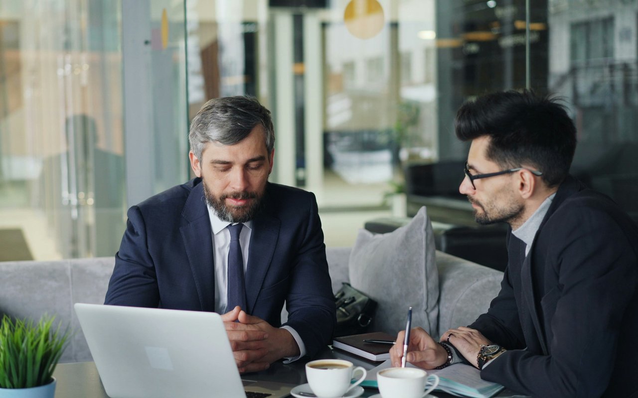 Two men discuss documents beside a laptop over coffee in a modern office.
