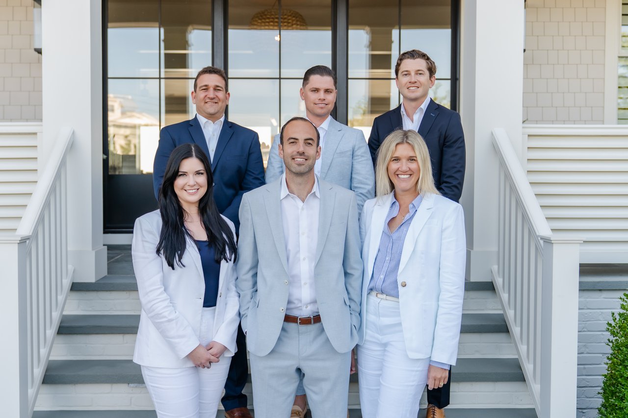 Six real estate agents pose together on the front steps of a house.