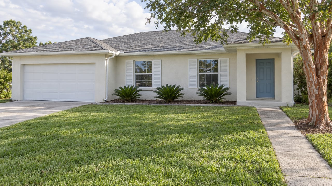 An ivory single-story home with a low shingled roof, white shutters and a broad lawn beneath a peeling copper-barked tree.