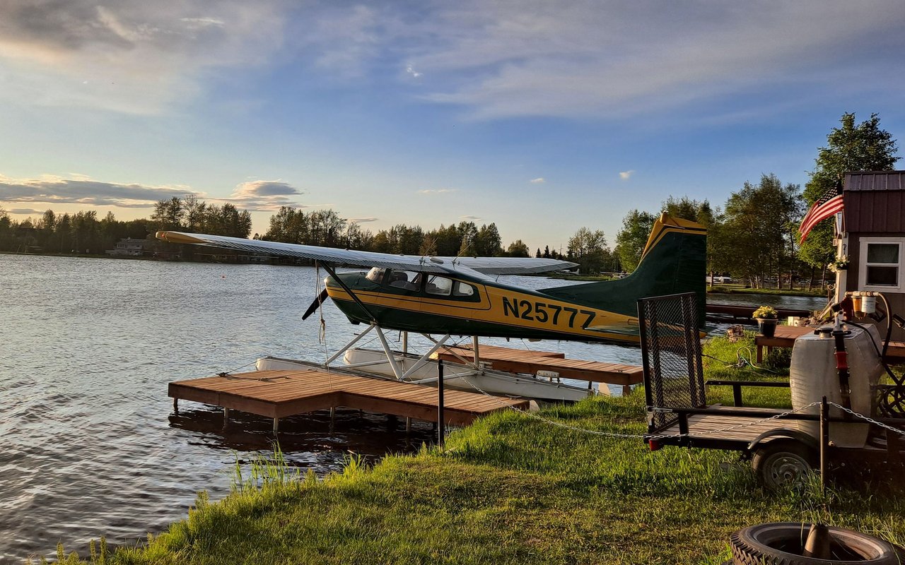Green-and-yellow floatplane moored at a lakeside dock beside a small building and American flag.