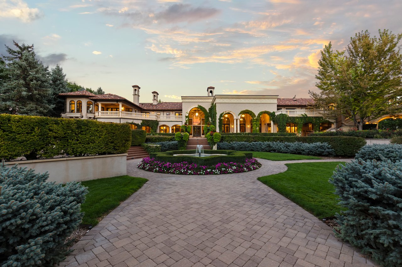 Estate grounds with a fountain, circular flower bed, clipped hedges, and Mediterranean-style home at sunset.