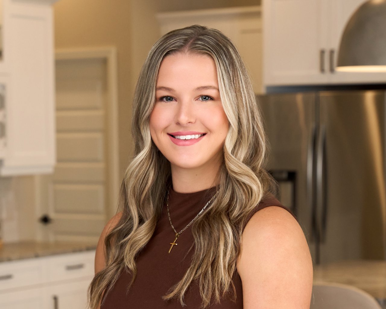 Maddie Cochran smiles in a kitchen, wearing a brown sleeveless top and cross necklace.