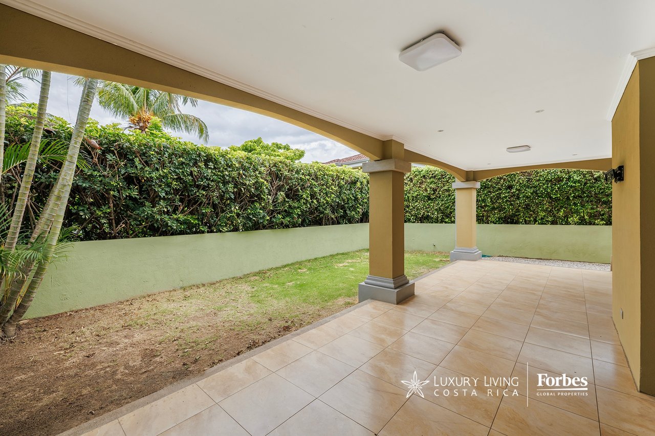 Covered tiled terrace beside a small garden, with palm trees and a tall hedge.