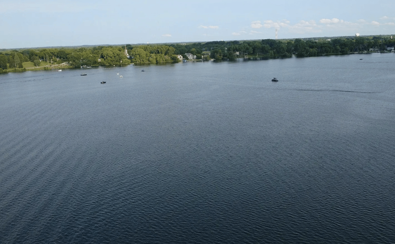 Aerial view across a lake toward a tree-lined shore with homes and boats.