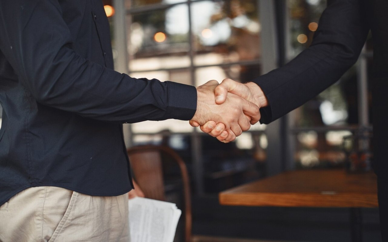 Two people shake hands across a table, with papers held by one person.