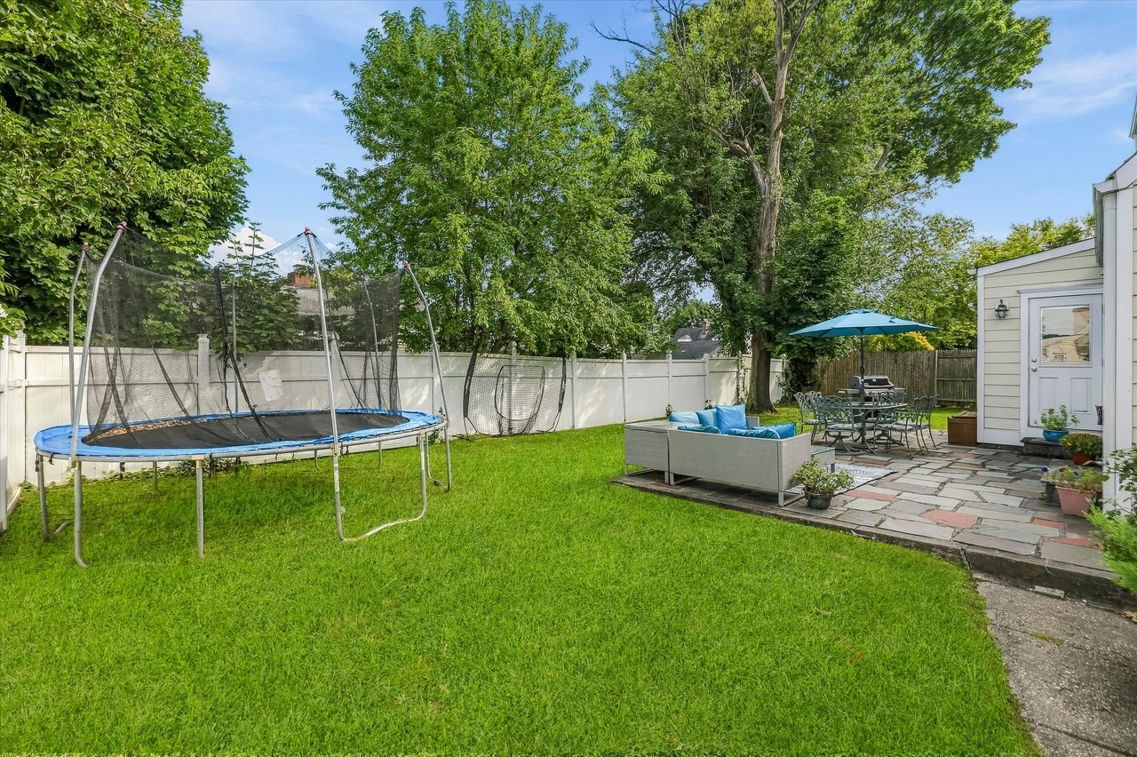 Fenced backyard with a trampoline, green lawn, and furnished stone patio beneath mature trees.