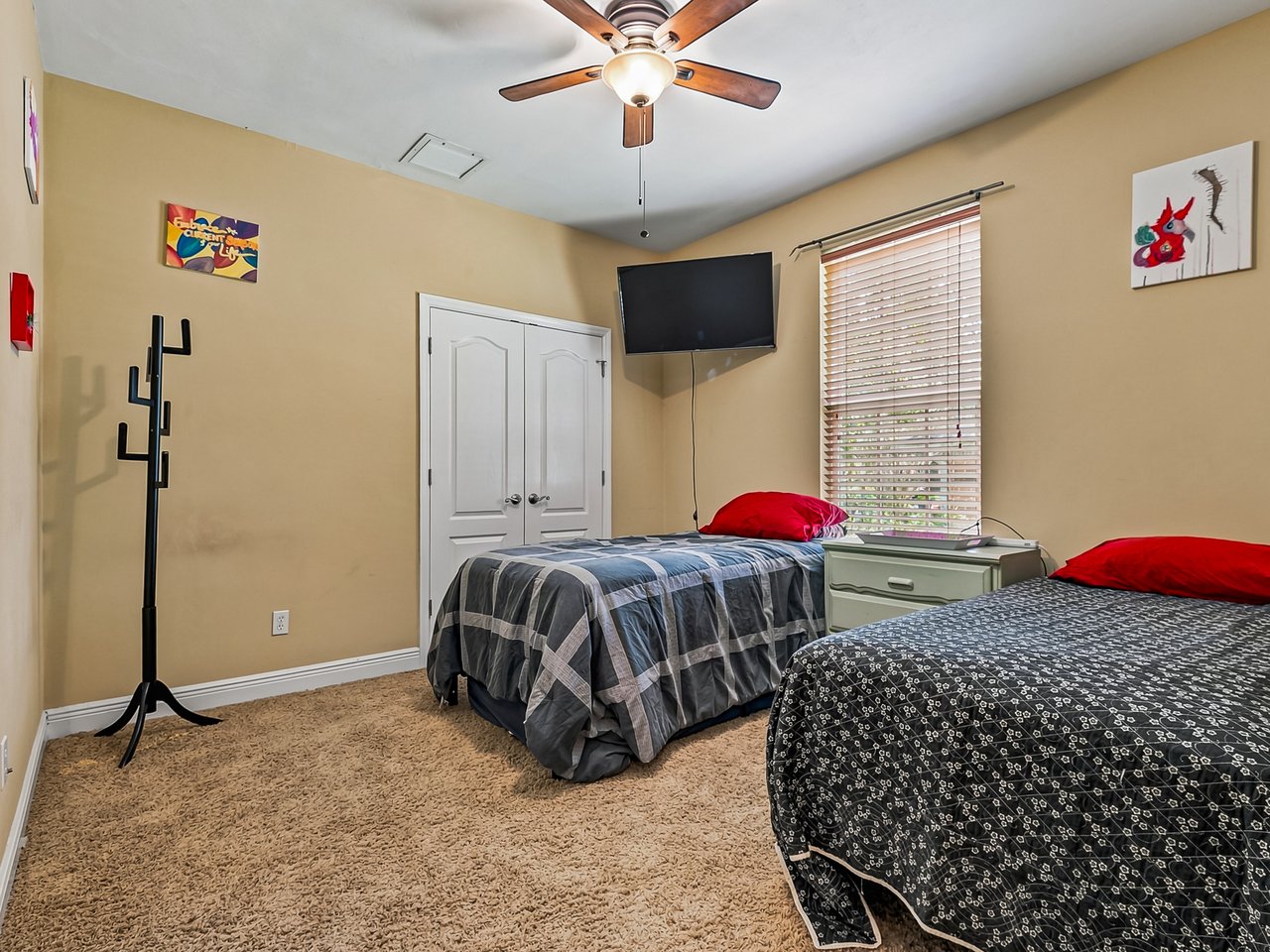 Furnished bedroom with two beds, beige walls, tan carpet, and a wall-mounted television.