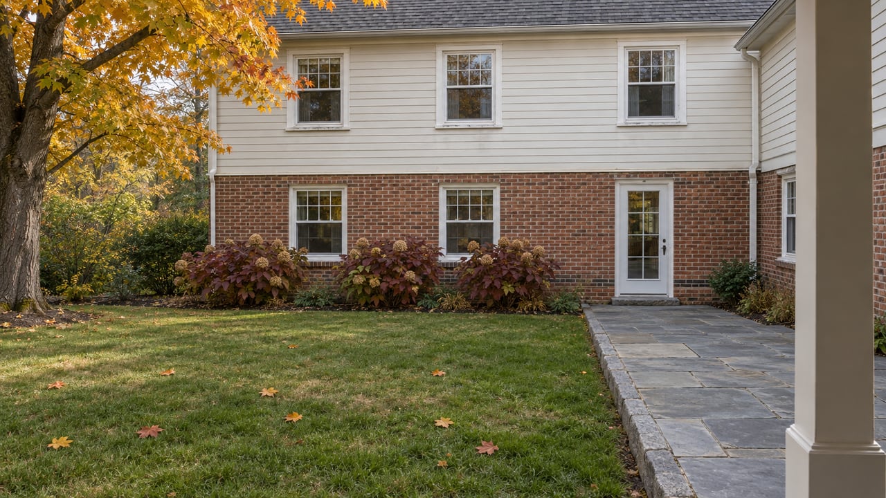 Autumn lawn and bluestone terrace beside a brick-and-clapboard home, framed from a covered rear porch.