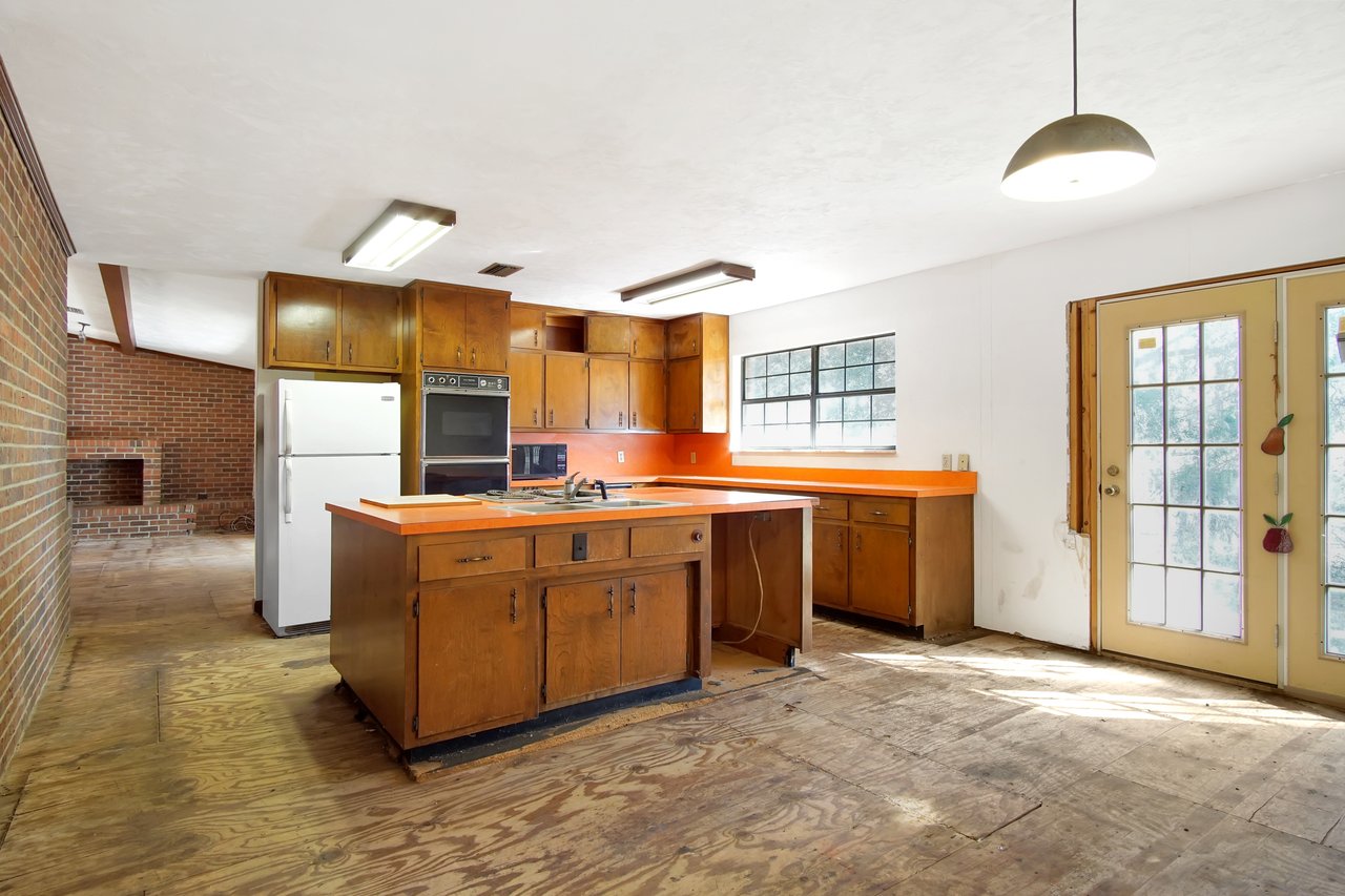 Retro kitchen with orange countertops, wood cabinets, and large windows. An exposed brick wall with a fireplace adds vintage charm. Bright natural light.