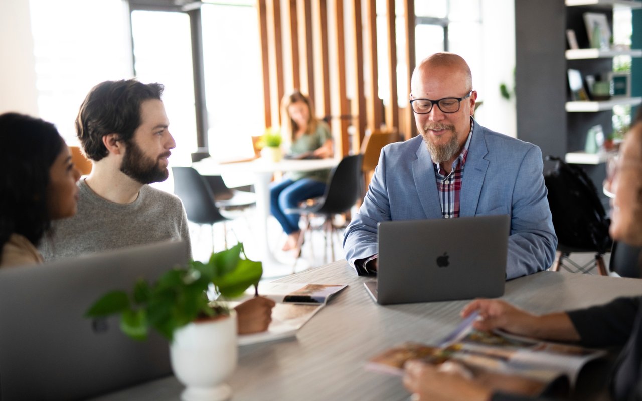 People discuss documents around a table as a man in a blue blazer works at a laptop.