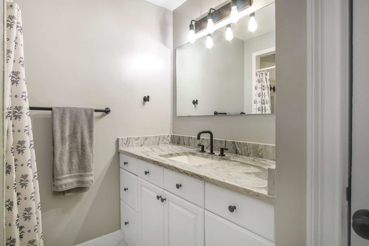 Modern bathroom with white cabinets, granite countertop, and large mirror. A towel hangs on a rod beside a patterned shower curtain. Bright lighting.