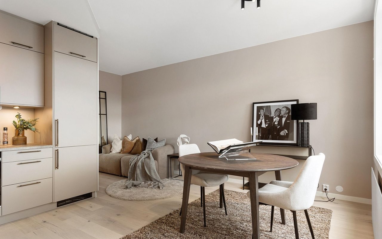 Open-plan dining area with a round wooden table, compact kitchen, and adjoining sofa nook.