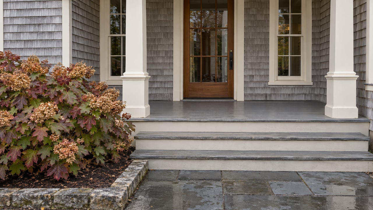 Rain-darkened bluestone steps lead to an ivory-painted porch and cedar-shingle entry, beside an oakleaf hydrangea with autumn foliage.