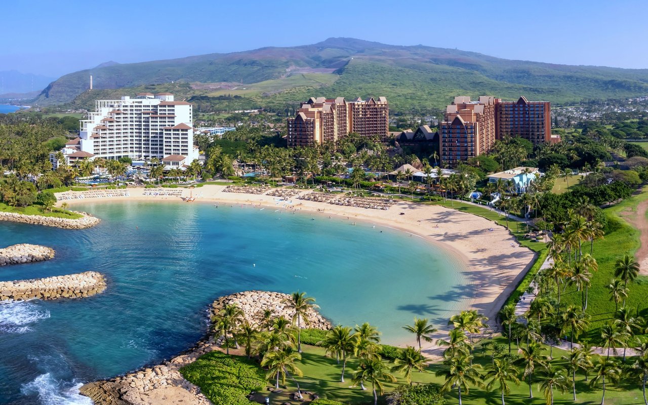 Curved sandy beach and turquoise lagoon beside palm-lined resort buildings, with green mountains beyond.