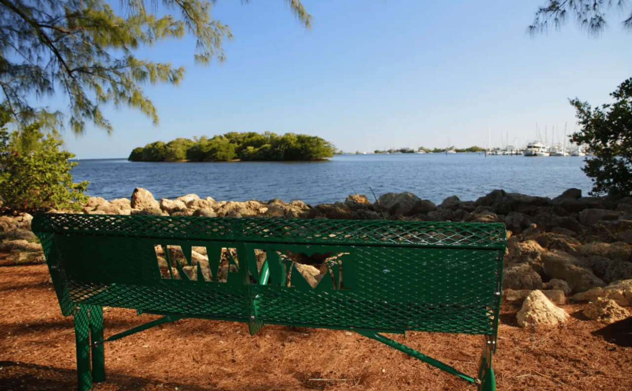 Green MIAMI bench beside the bay, with a mangrove island and boats in the distance.