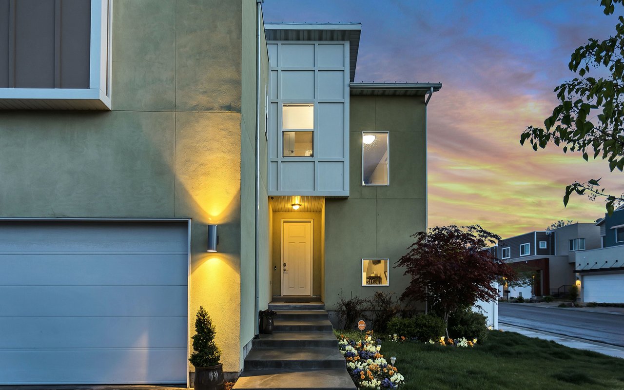 Modern multi-story home at dusk with a lit entry, attached garage, and colorful flower beds.