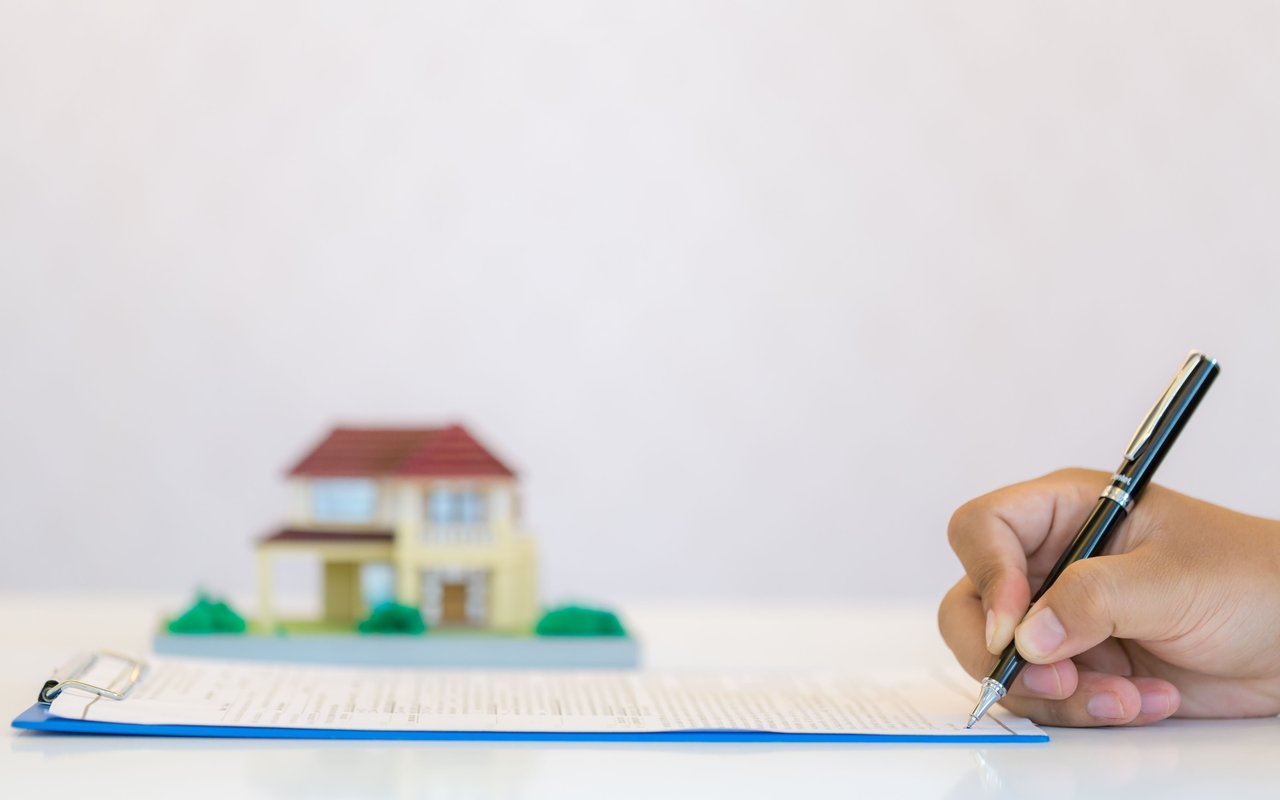 A hand signs mortgage paperwork beside a small model house.
