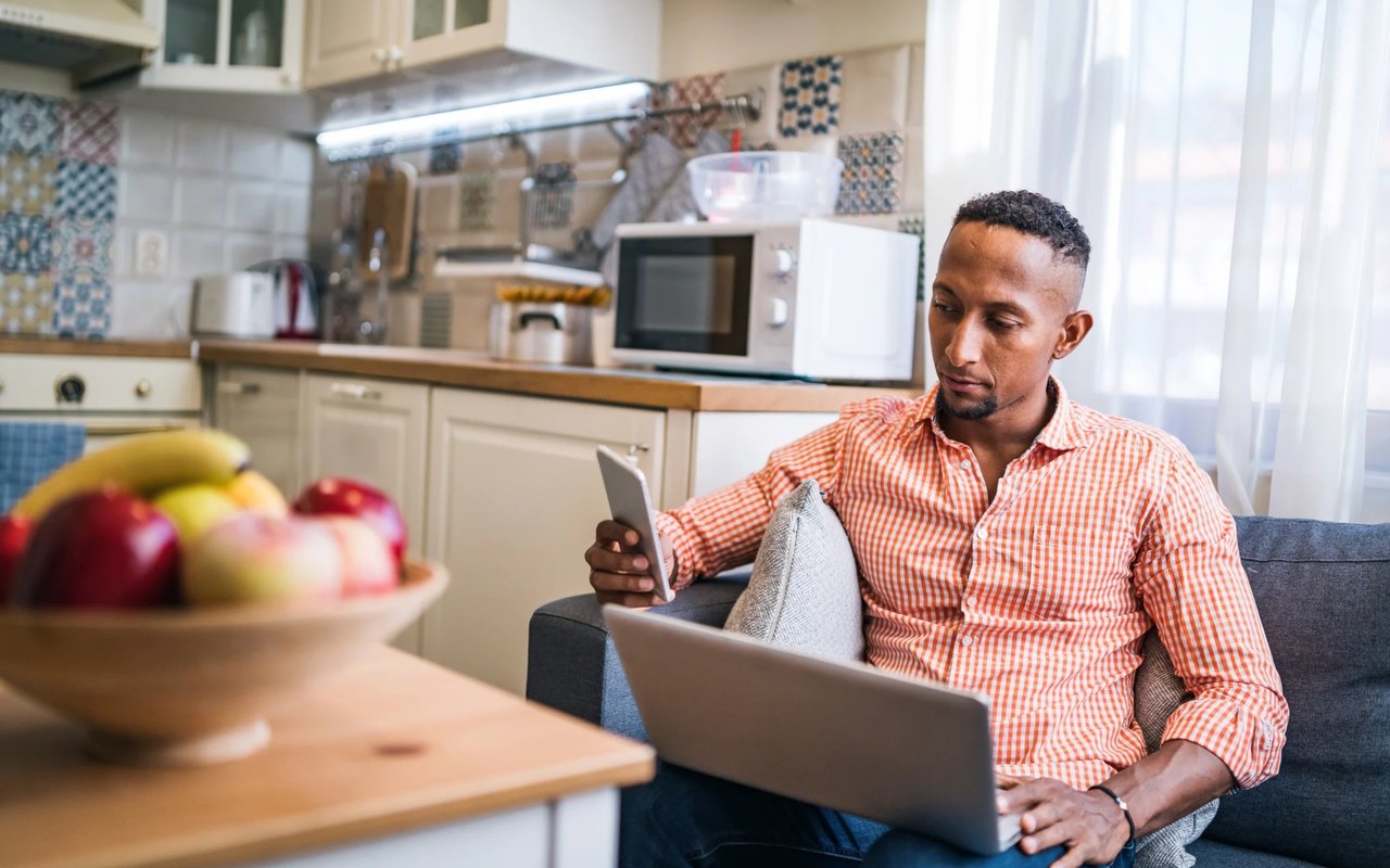 Man checks his smartphone while using a laptop on a sofa in a bright kitchen.