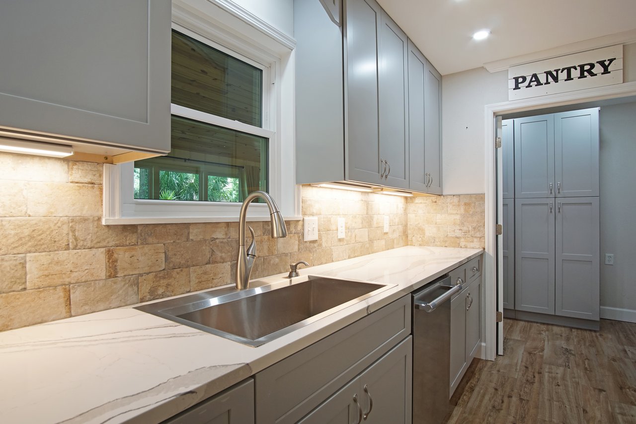 Modern kitchen with gray cabinets, marble counter, stainless steel sink, and dishwasher. Warm tone lighting, stone backsplash, and pantry door.