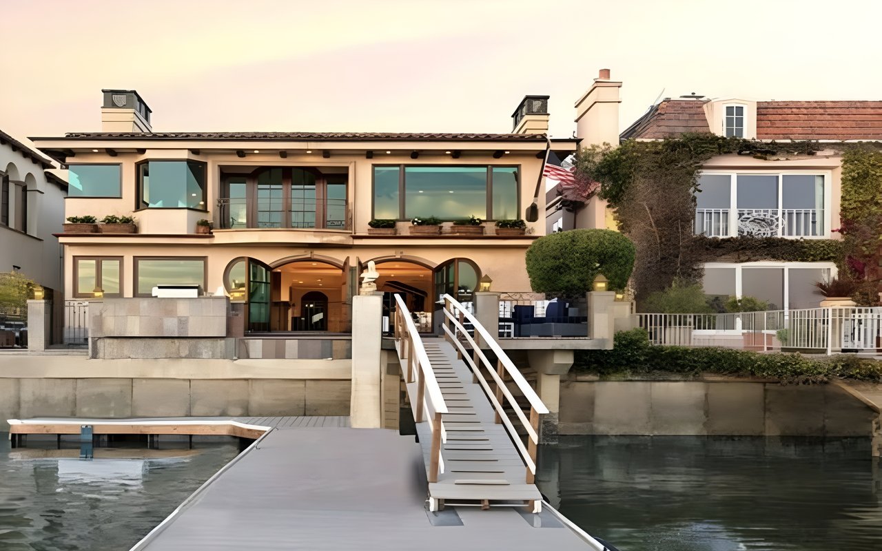 A two-story waterfront house in Newport Beach with a private dock leading to the water, under a warm, golden-hour sky.