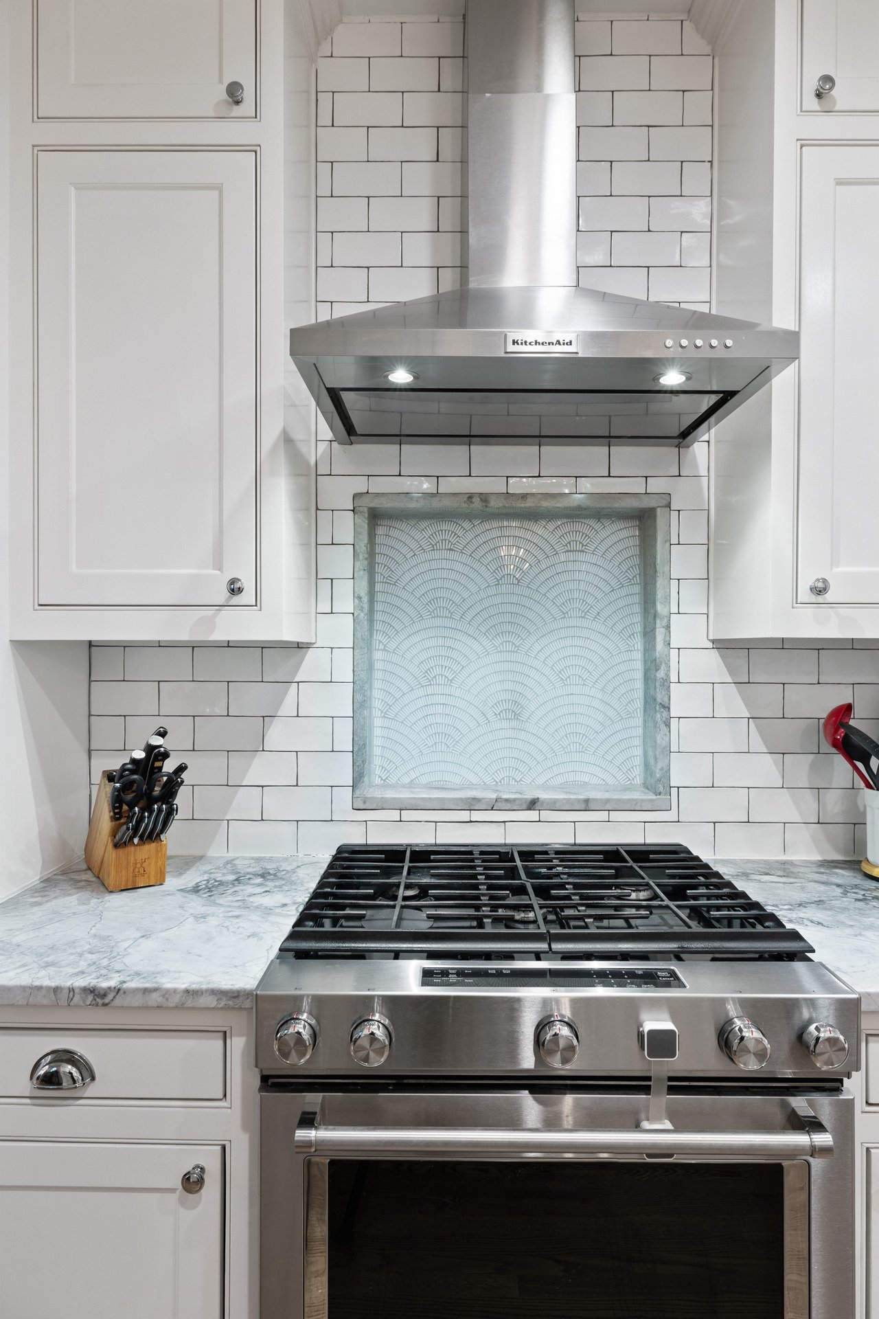 Kitchen range beneath a stainless-steel hood, with a patterned tile inset and marble countertops.