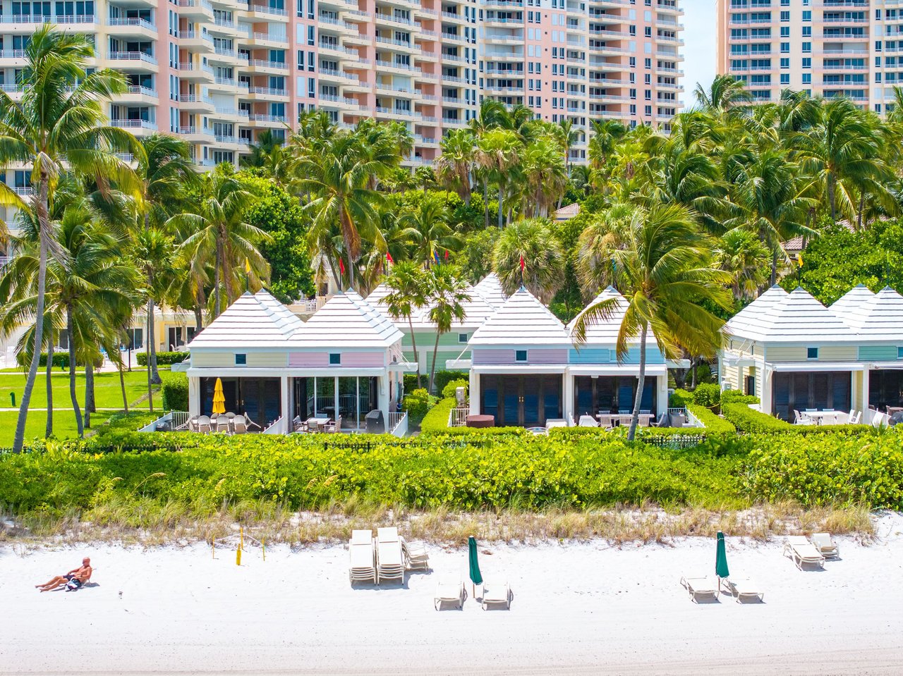 Beachfront cabanas beneath palm trees, with a pink-and-white resort tower behind them.