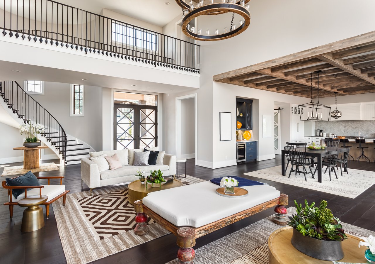 Open-plan living room with a curved staircase, upper balcony, and kitchen beneath a timber ceiling.