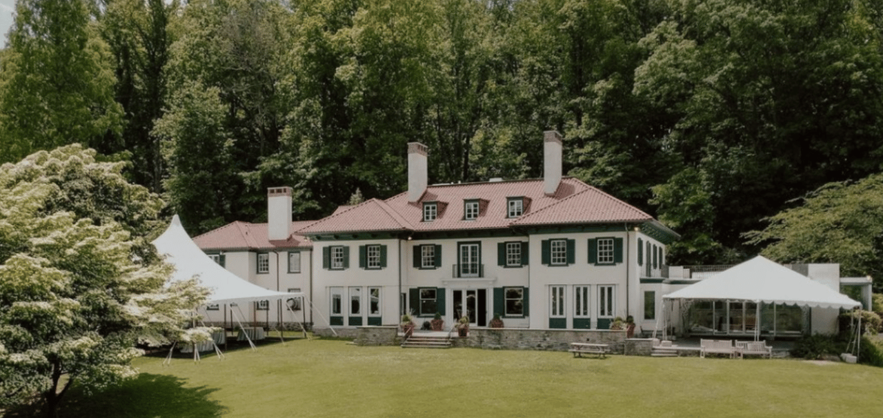 Large white estate house with green shutters, red tile roof, broad lawn, and white event tents.
