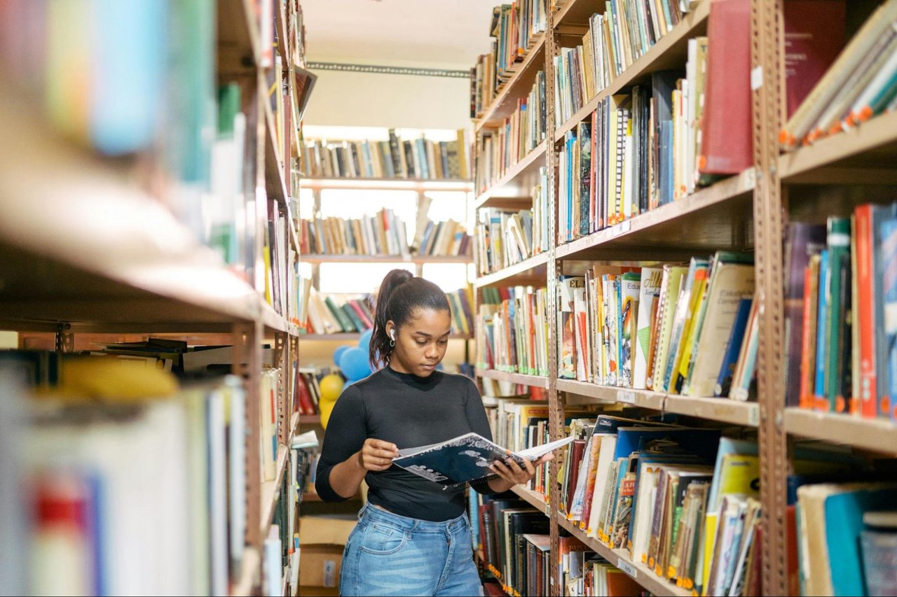 Woman browsing a magazine between tall shelves filled with books.