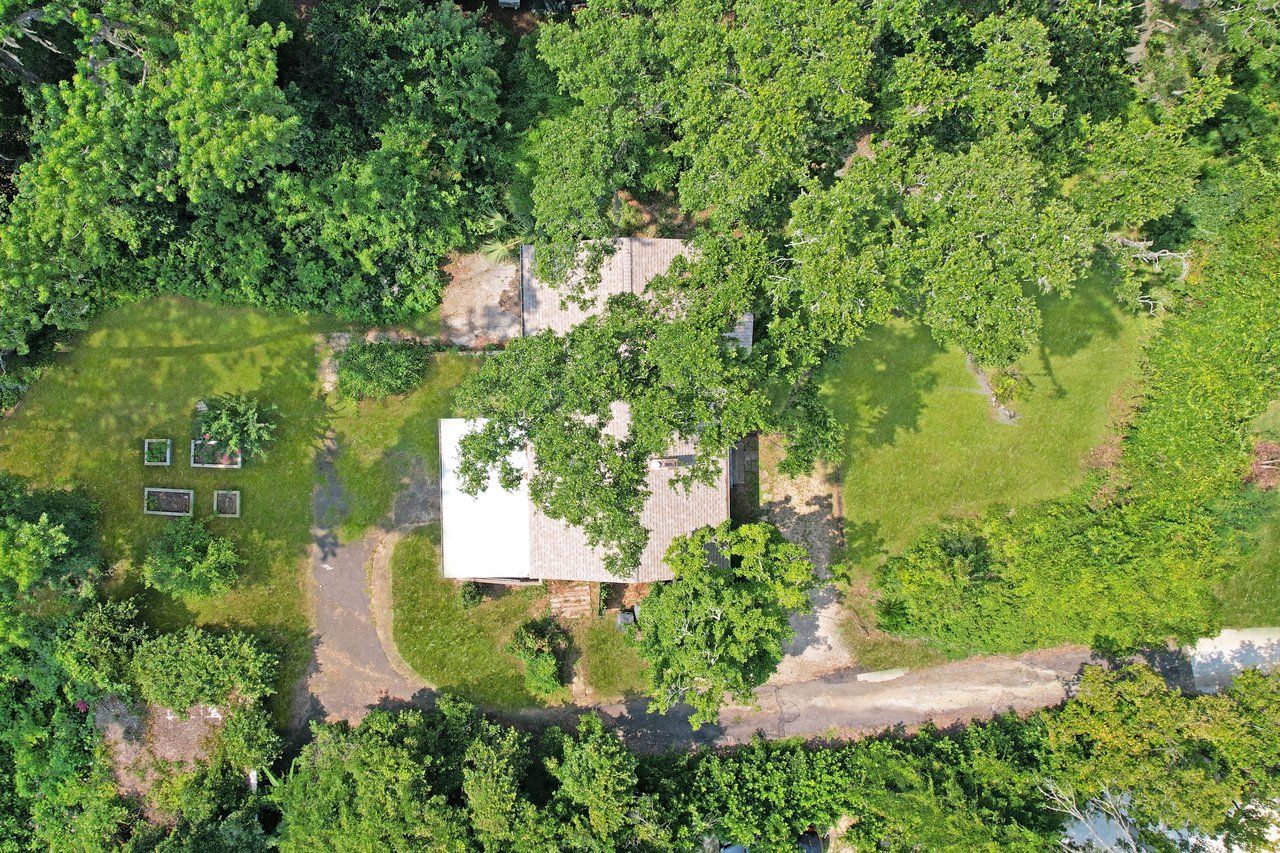 Aerial view of a house surrounded by lush green trees and grass. A driveway curves in front, with a garden featuring raised beds on the left, creating a serene, natural atmosphere.