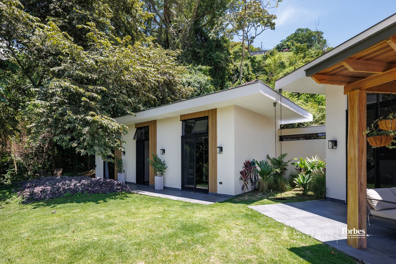 Modern home with white walls, wood accents, large windows, and a landscaped lawn beneath tropical trees.