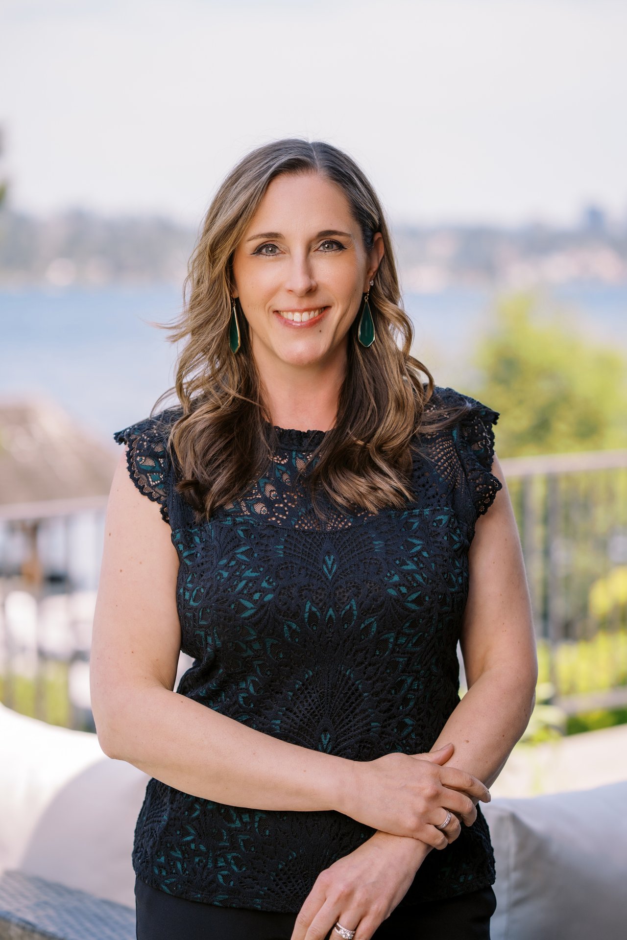 Headshot of professional real estate agent smiling outdoors with waterfront background, wearing a black lace top and green earrings.